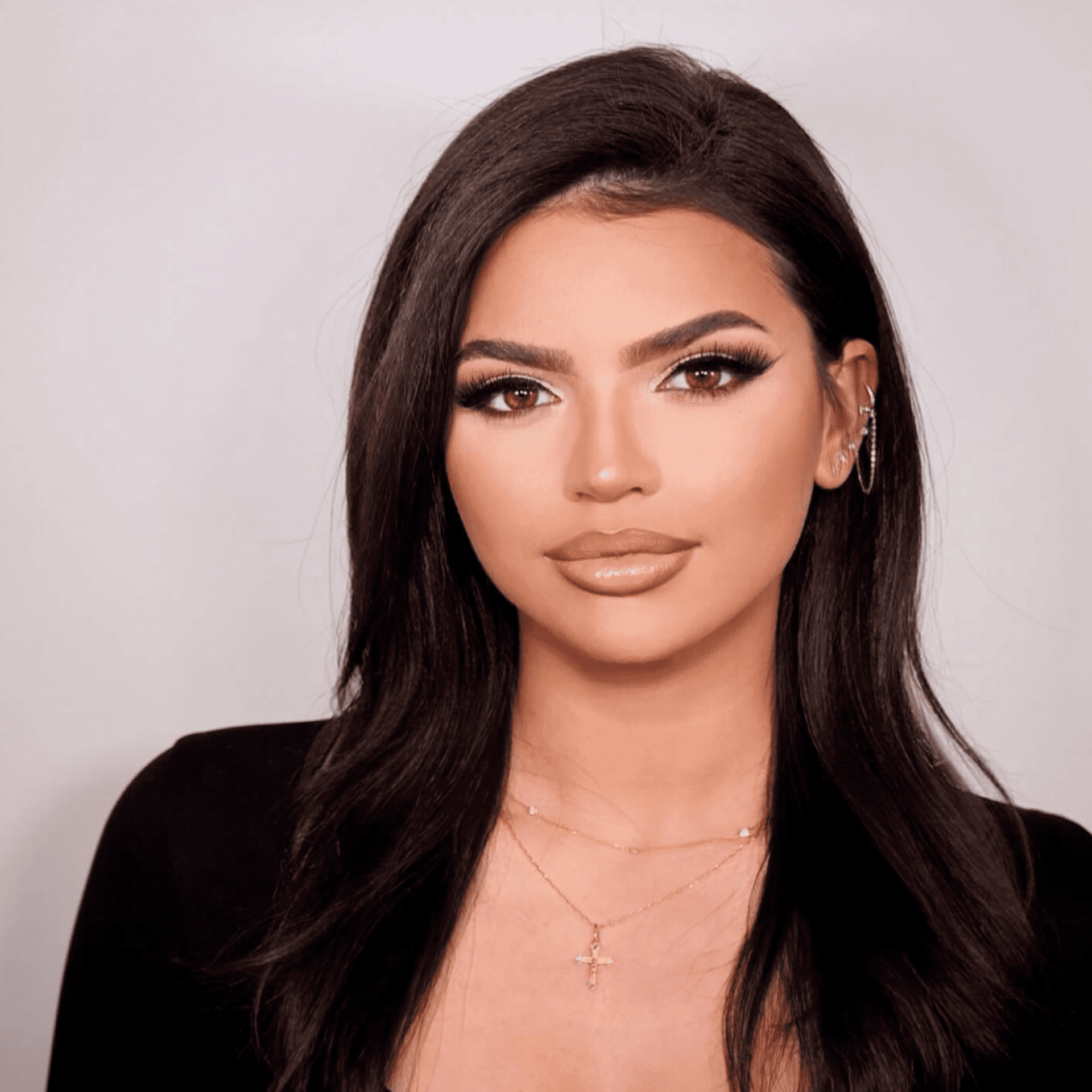 Woman with dark hair, wearing makeup and jewelry, against a light background.